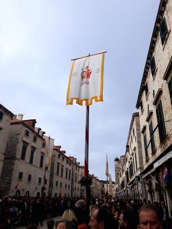 Bandera blanca y dorada con la figura de San Blas, en lo alto de un mástil, en una calle de Dubrovnik llena de gente