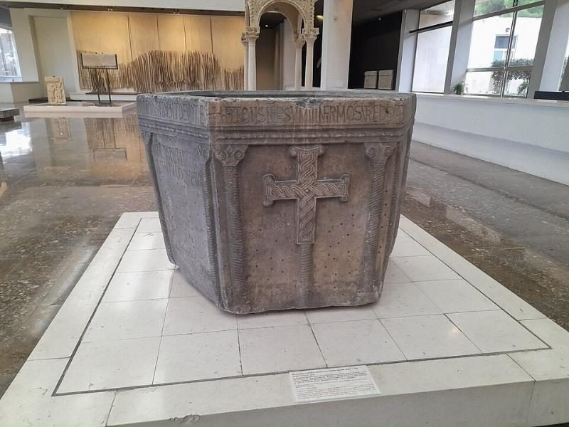 Pila bautismal de piedra con una cruz tallada, exhibida en el centro de una sala de museo