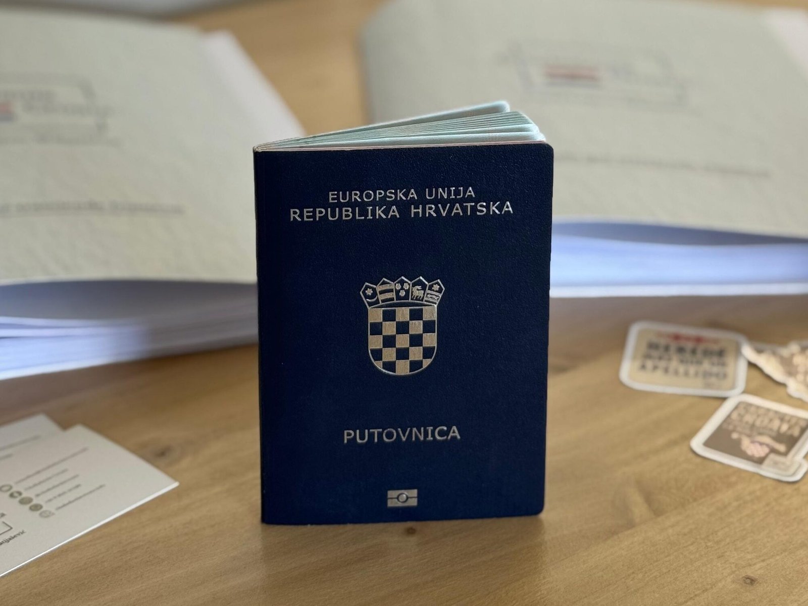 Pasaporte croata de pie sobre una mesa de madera, con carpetas de documentos abiertas detrás y tarjetas y stickers de Ciudadanía Croata alrededor